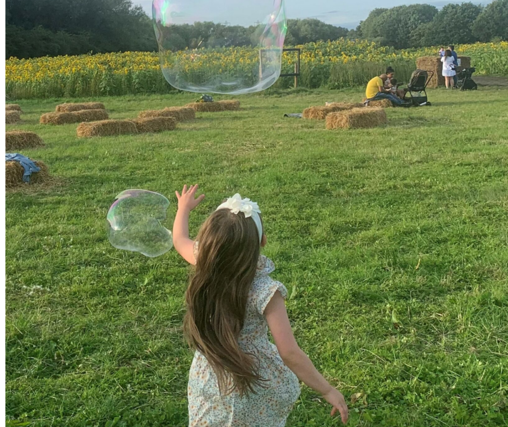 A little girl joyfully plays with bubbles in a sunlit field surrounded by sunflowers at sunset.